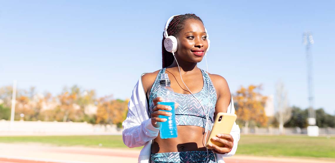 A woman standing on an outdoor track while wearing headphones and holding her phone and an energy drink.