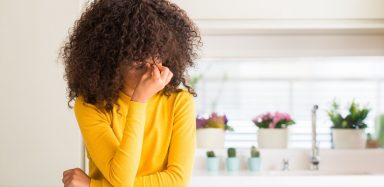 A woman with curly brown hair is leaning on a counter, holding her face in her hand. She is wearing a bright yellow shirt.